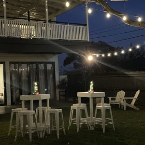 Warm-white festoon lights strung across a backyard at dusk over white bar tables and stools